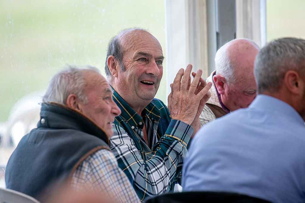 A table of older gentlemen dressed in country attire smile at something off camera.