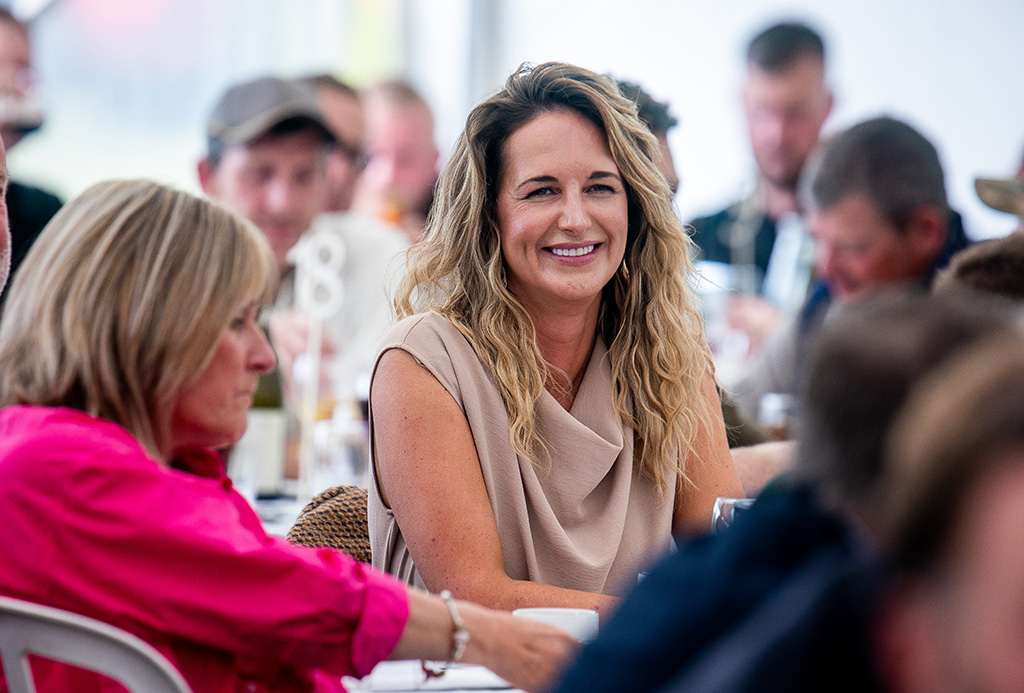 A smartly-dressed white woman in her forties with long wavy hair smiles at someone in the distance,