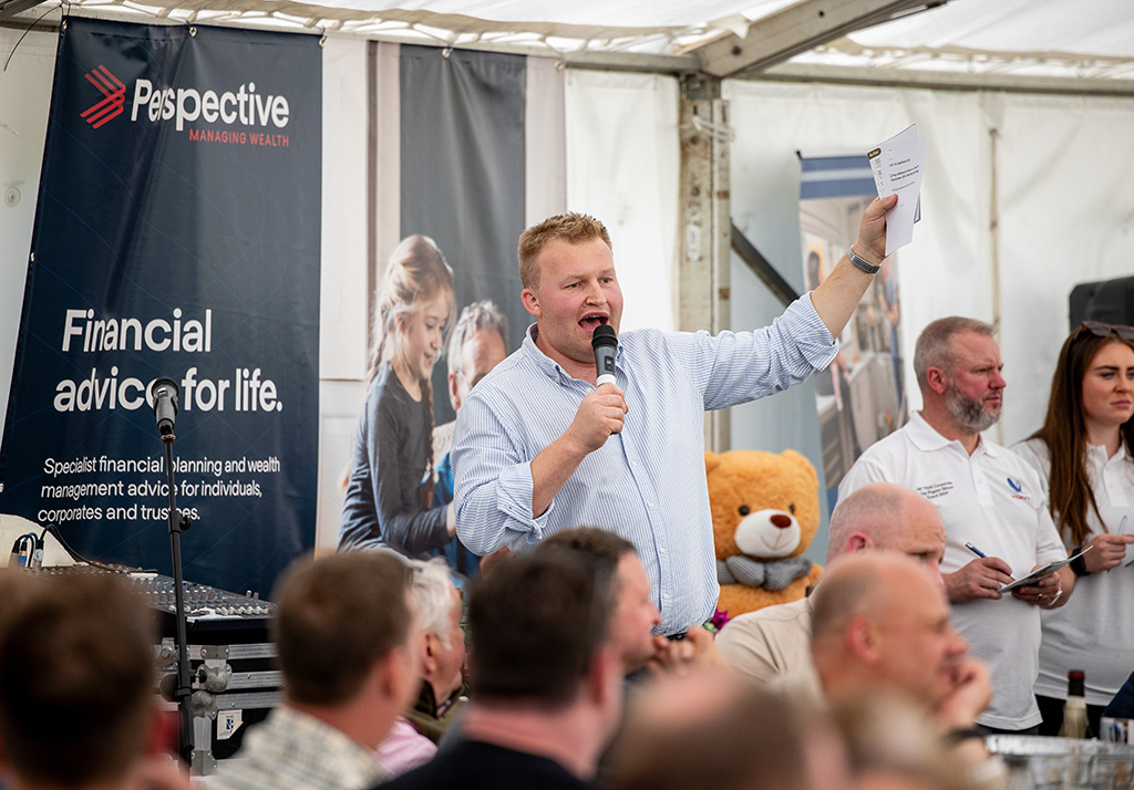 A white male, well built in a light striped shirt, holds aloft a programme as he speaks to a room full of people through a microphone. He is an auctioneer.