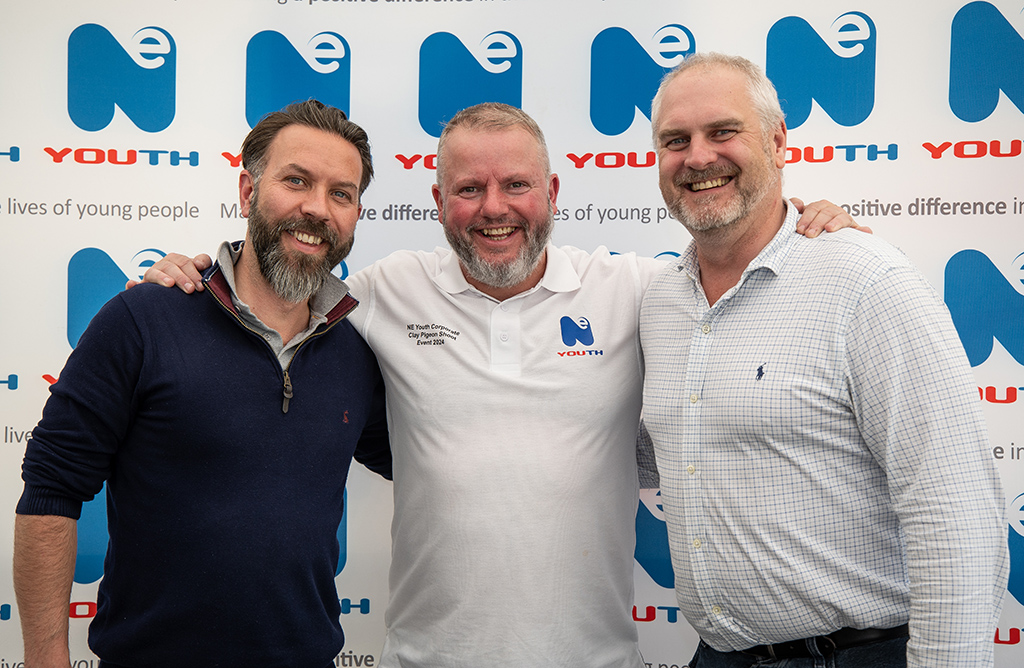 Three white middle-aged men stand in front of a branded backdrop covered entirely by NE Youth logos. The men are smiling, happy and have arms around each other's backs. They are dressed casually in shirts.