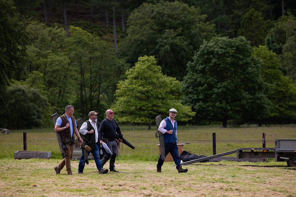 Four white men visible in the distance walk in a field. They are carrying shotguns in their cases.