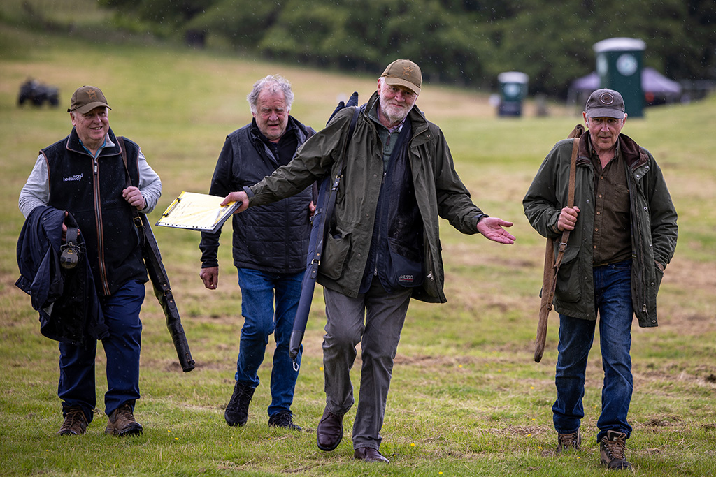 Four older men walk back from a clay pigeon shoot dressed in country attire such as Barbour jackets. They are in a countryside setting, carrying coats and shotguns. One has his arms outstretched.