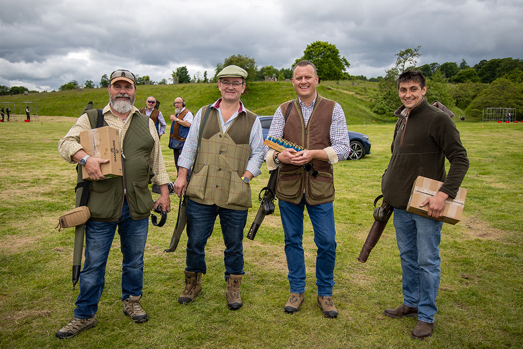 Four white men stand in a row, holding shotguns and cartridges as they prepare to shoot clay pigeons in a green countryside setting.