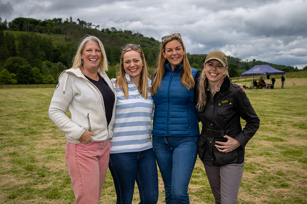 Four women stand in a row, smiling at the camera. They are in a green countryside setting.