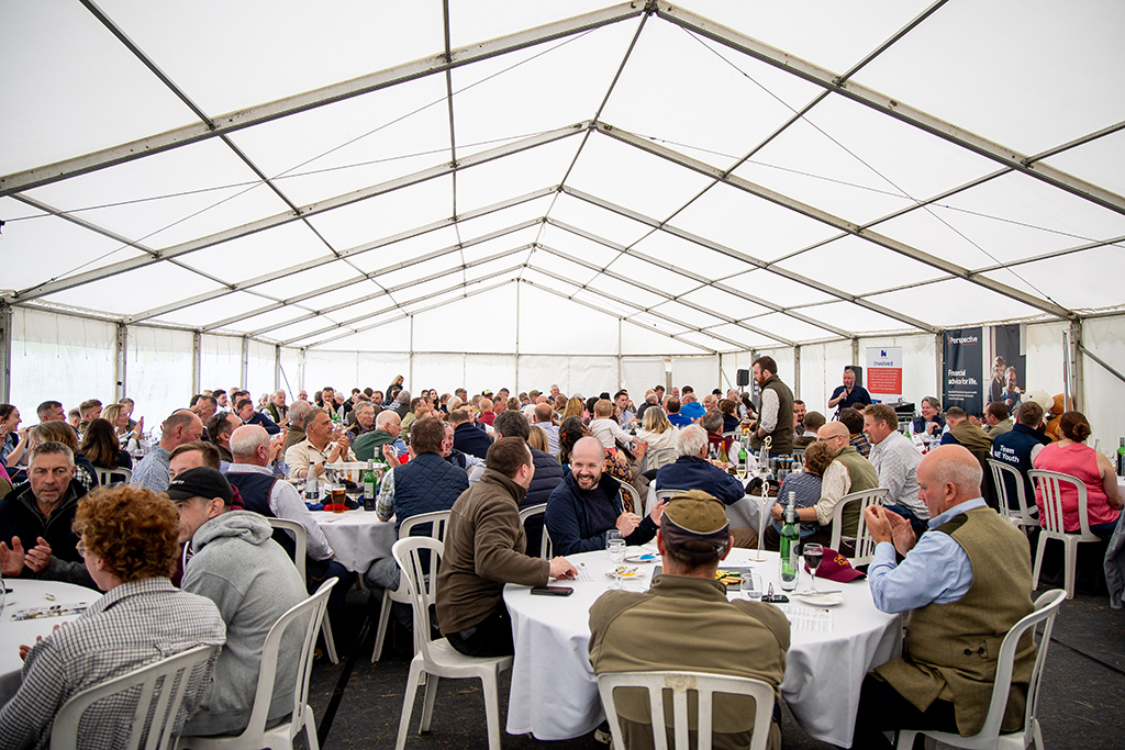 Around 200 people are seated in a large white marquee tent. They are mostly white and in middle age. They are seated at round tables with food and drinks visible.