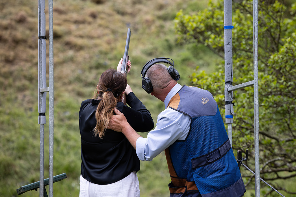 A woman points a shotgun upwards as she takes aim at a clay pigeon. An official hold her styeady as she is about to shoot.