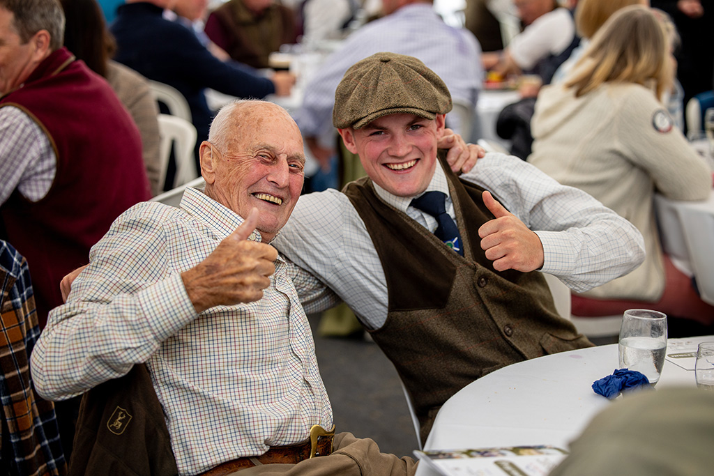 Two white men, one young and one old, give the camera a thumbs-up and a smile as they sit at a table.