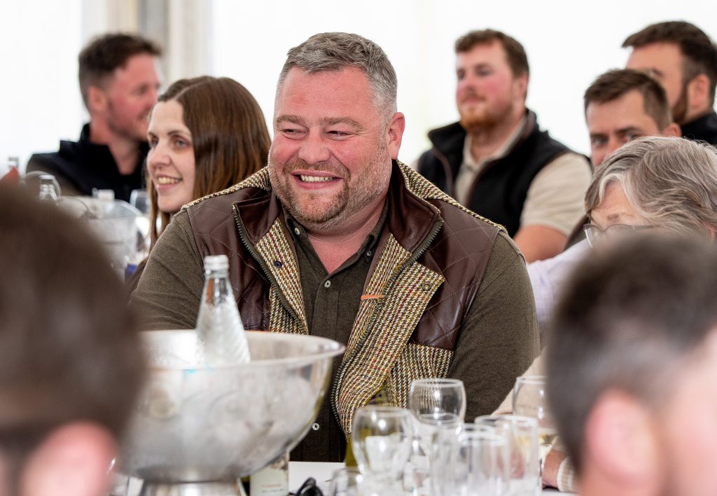 A white man with short grey hair laughs at something off camera as he sits at an event surrounded by people.