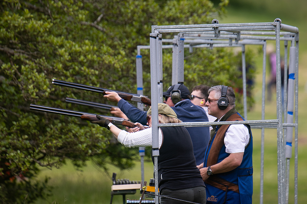 A group of people aim shotguns in a dedicated shooting trap. The guns are pointed slightly upwards, ready to fire. They people are dressed in countryside attire, and all wear ear protectors. The backdrop is English countryside.