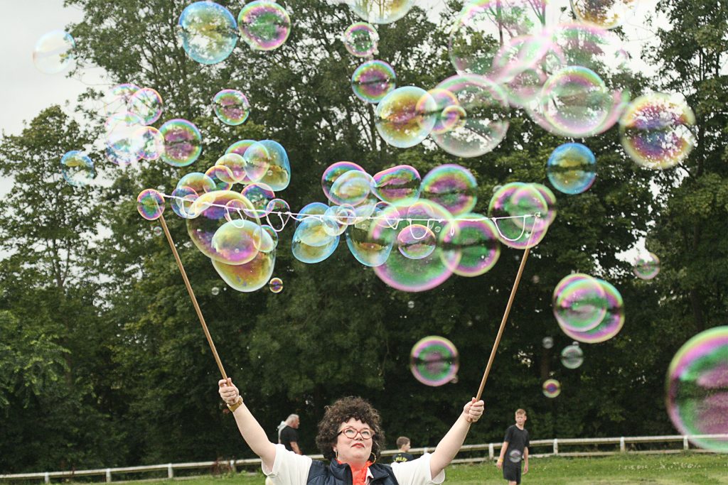 A white woman with curly dark hair and glasses holds two sticks aloft, generating dozens of soap bubbles that fly into the air.