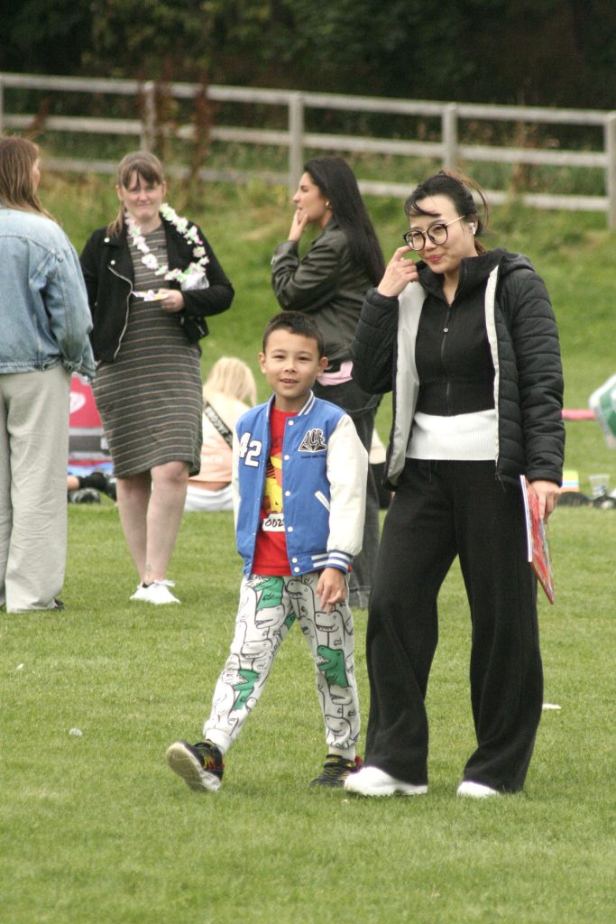 An East Asian mother and child walk across a field together in conversation. His clothing has cartoons visible, the mother all in black with long hair and glasses.