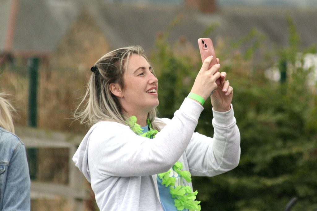 A white woman in her mid-twenties looks upwards, holding a smartphone aloft as she photographs something out of shot. She has a bright garland and green bracelet.