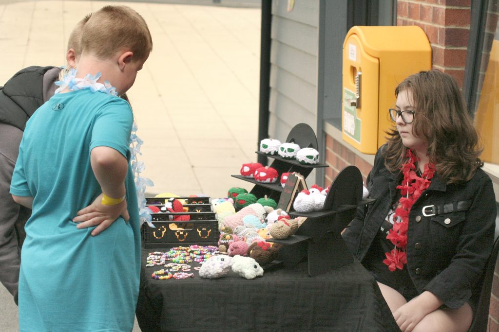 A young white boy peruses a choice of hand-crafted crocheted animals on an outdoor stall. A white teenage girl with dark hair and glasses is seated, looking on.