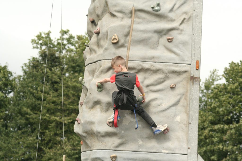 A young white boy with short mousy hair climbs an artificial climbing wall outdoors. His security harness and ropes are visible, as the smiling child looks downwards.