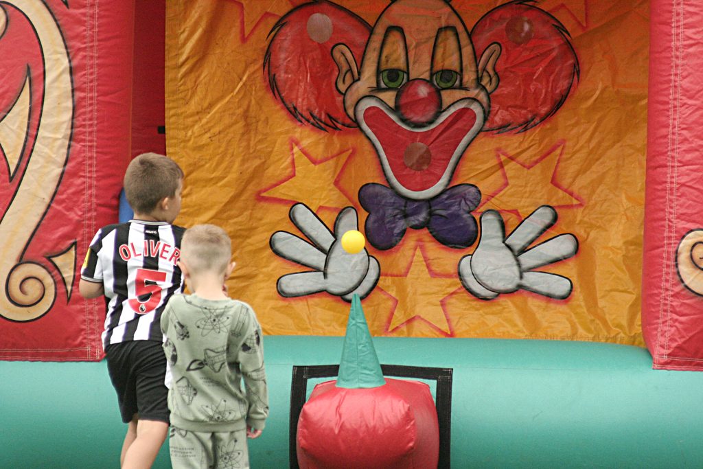 Two young white boys, one dressed in a football kit, play a bat and ball game with the backdrop of a large, bright, inflatable structure featuring a clown.