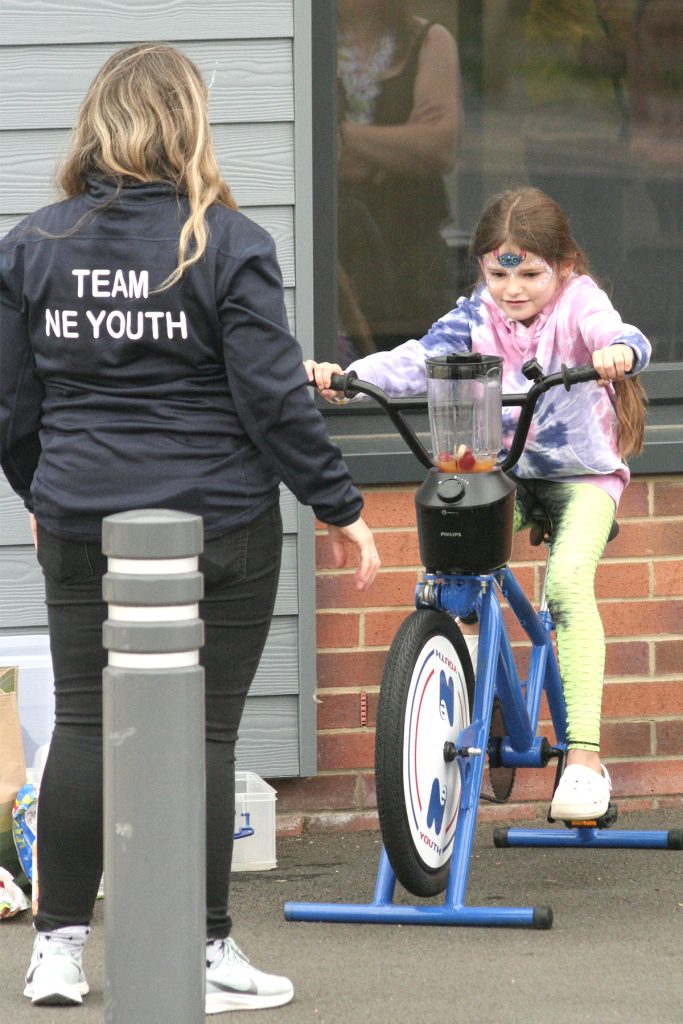 A young girl, white, with long dark hair and dressed in bright tie-dye clothes pedals on a smoothie bike to blend her fruit into a drink. An adult is visible from the rear as she looks on.