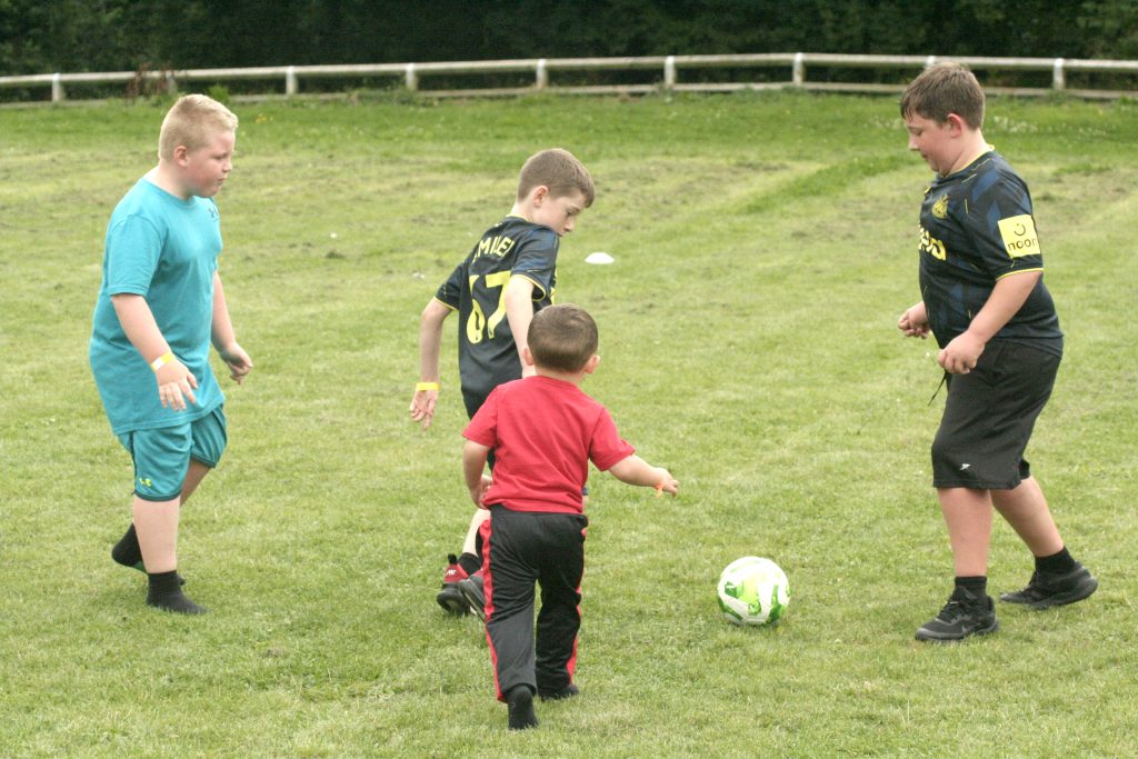 Four white biys of various ages play football on a grass field. Two are wearing football shirts, the smallest bout wearing a red t-shirt.