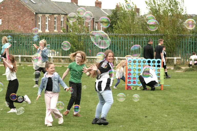 Children laugh as they chase giant bubbles at an outdoor community event.