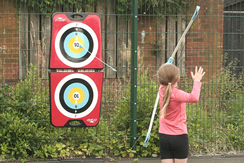 A young girl with fair hair shoots an arrow with a sucker at the end towards a target, hitting the bullseye.