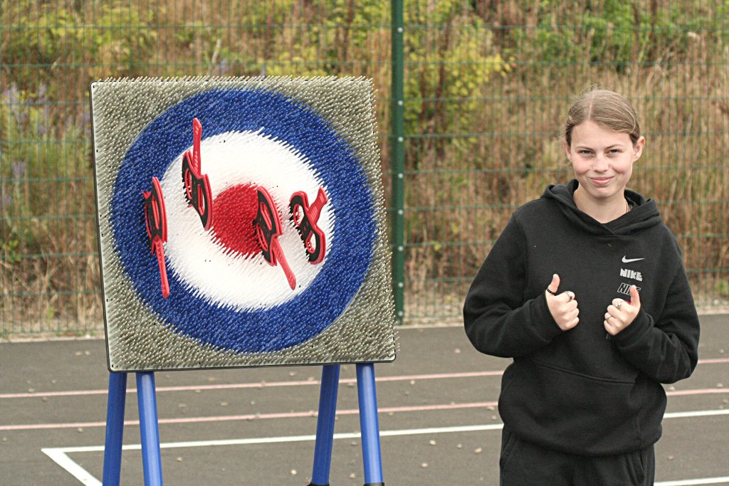 A white teen girl stands next to a large target with plastic axes embedded in it. One axe is in the bullseye. She is looking at the camera with both hands in a thumbs up position. She is dresed all in black, with fair hair in a ponytail.