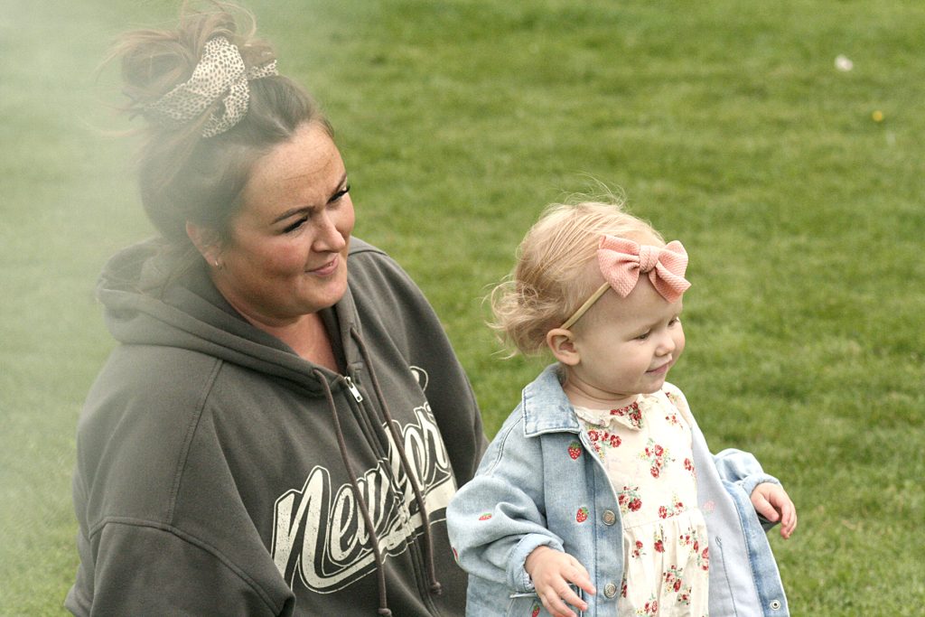 A white mother and toddler daughter look on as other children play. The toddler has a bow in her hair and a flowery dress. The mother wears a hoody and has her brown hair tied up.