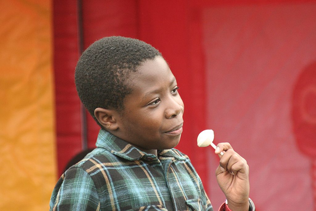 A close-up of black boy around 11 years old stands with a lolipop agains a bright red background. He is half smiling with his head at a slight angle.