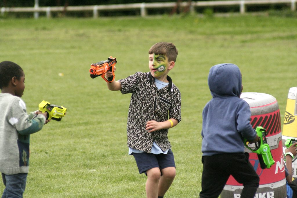 Three boys energetically play with Nerf guns outdoors, two black and one white. The white boy has Hulk face paint and a patterned shirt. One boy has his hood up, and the other is looking down at his gun.