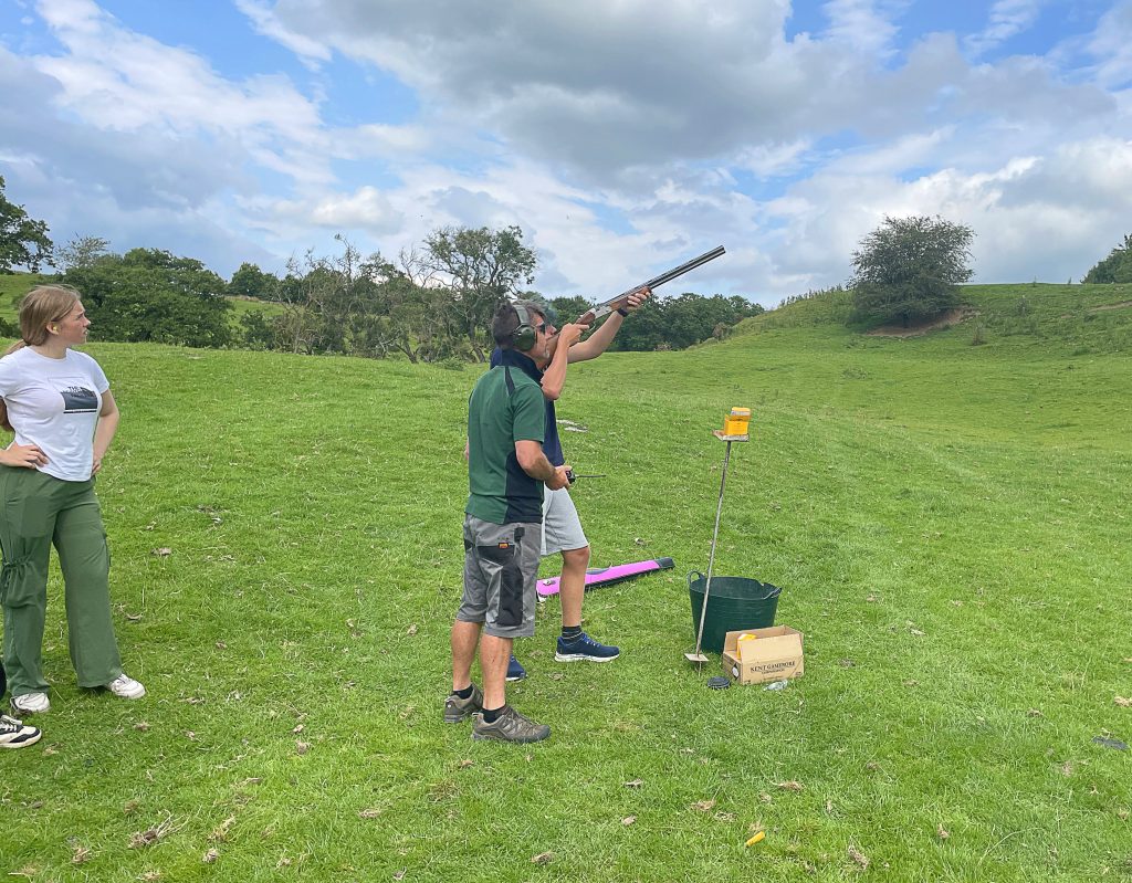 A teen boy aims a shotgun at a clay pigeon preparing to fire under the watchful eye of an adult. They are in the countryside.