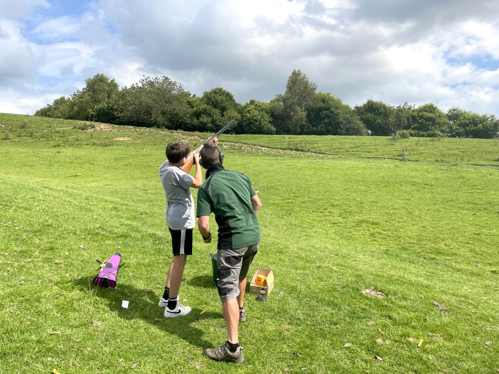 Teen boy points shotgun to the sky under the supervision of an adult . They are in a countryside setting with a dramatic cloudy sky.