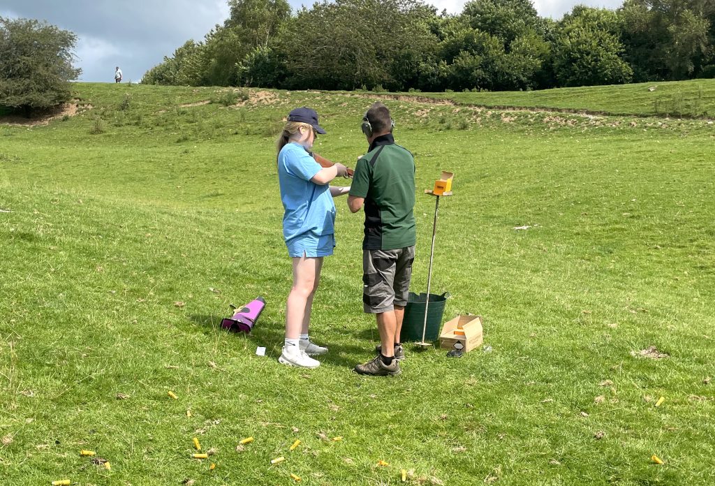 A teen girl loads her shotgun at a clay pigeon shoot event. An adult male instructor is supervising in the British countryside.