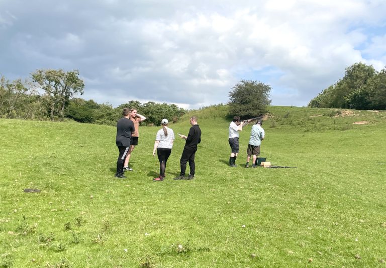 A group of teens stand by as a peer aims his shotgun towards a clay pigeon. Countryside setting.