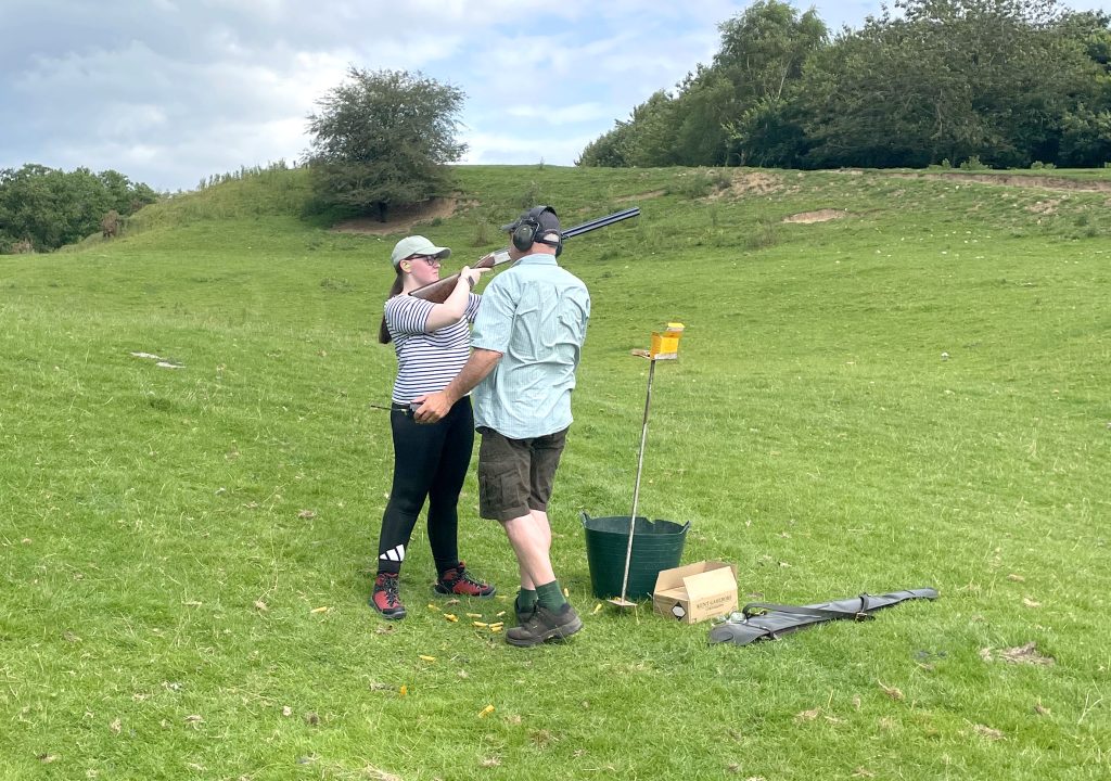 A teen girl prepares to shoot her shotgun at a clay pigeon, supervised by an adult.