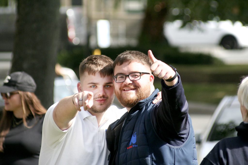 Two young men look directly into the camera, pointing and giving a thumbs-up. Both white, bearded men smile. One is wearing glasses.