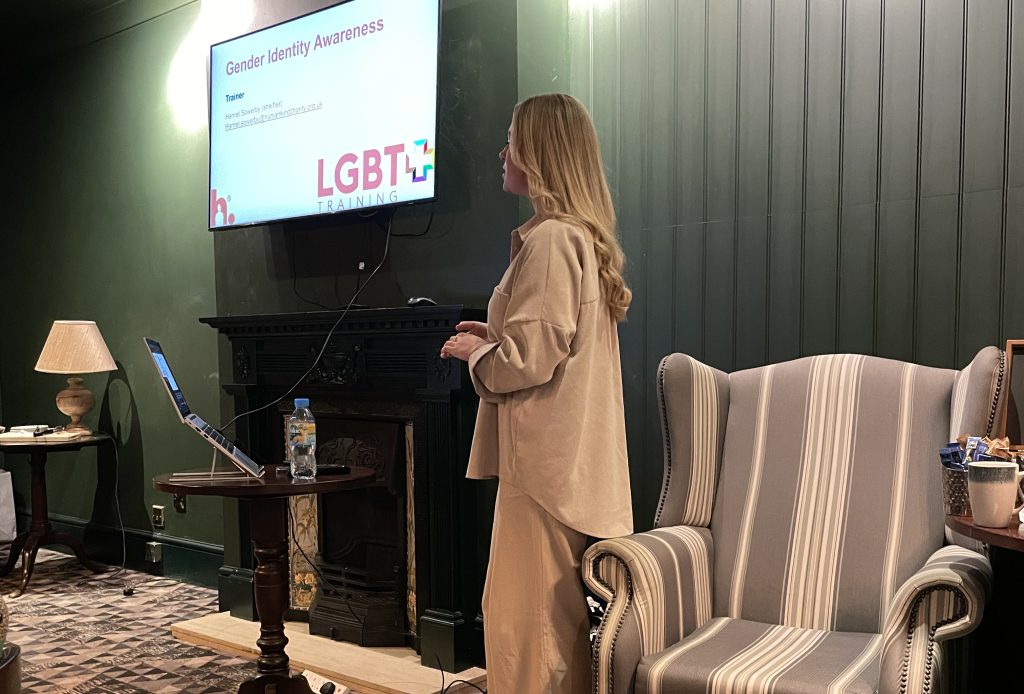 A female speaker dressed in loose beige clothing stands as she delivers a seminar. She stands next to a striped wing-back armchair and Victorian fireplace. A lit screen is above her, linked to a laptop on an adjacent table.