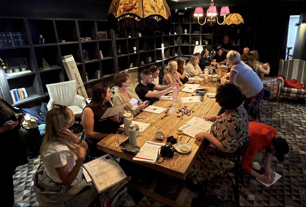 A group of people are sat at a long wooden table, in discussion as they look at folders in front of them in a moodily-lit room bordered by bookshelves with books and other items.