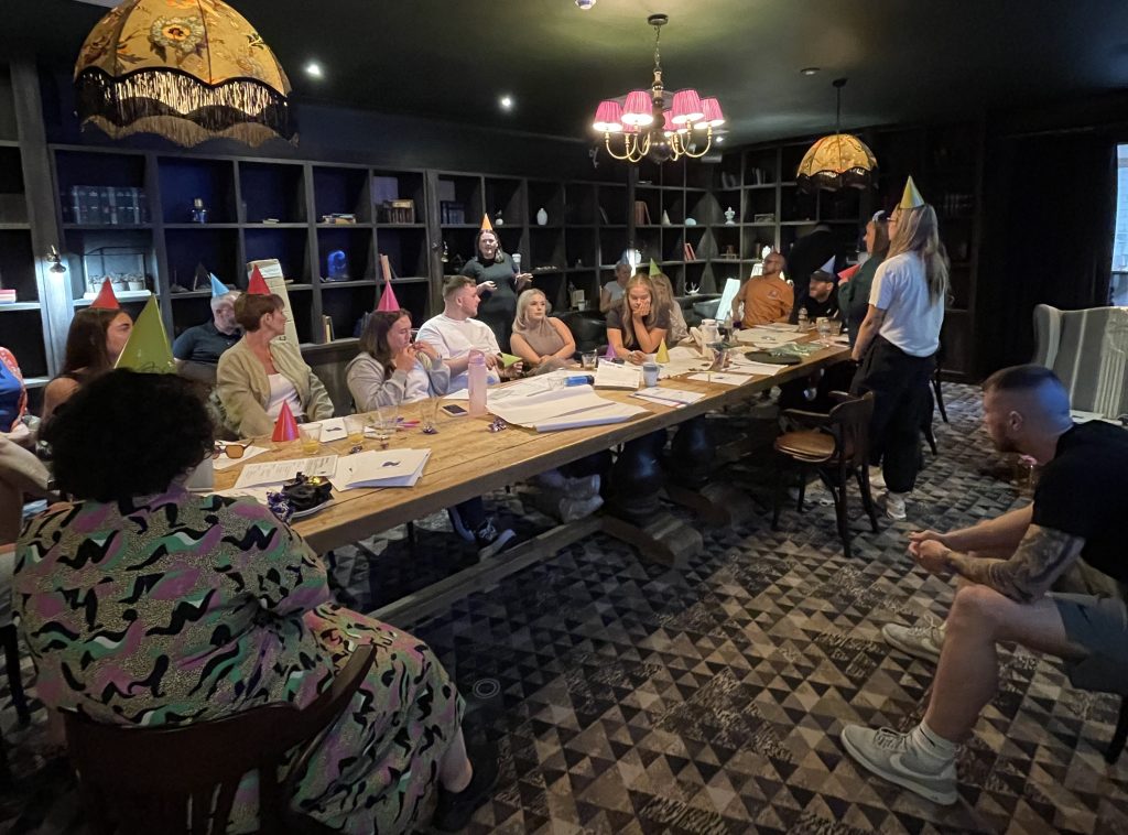 A group of people are sat at a long wooden table, listening intently to a speaker. They are wearing pointed party hats as part of a team-building task.