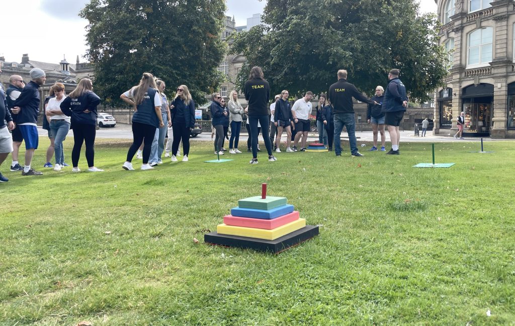 A group of colleagues stand together on a patch of grass in Harrogate, England. They await the beginning of outdoor team-building games, dressed in NE Youth clothing.