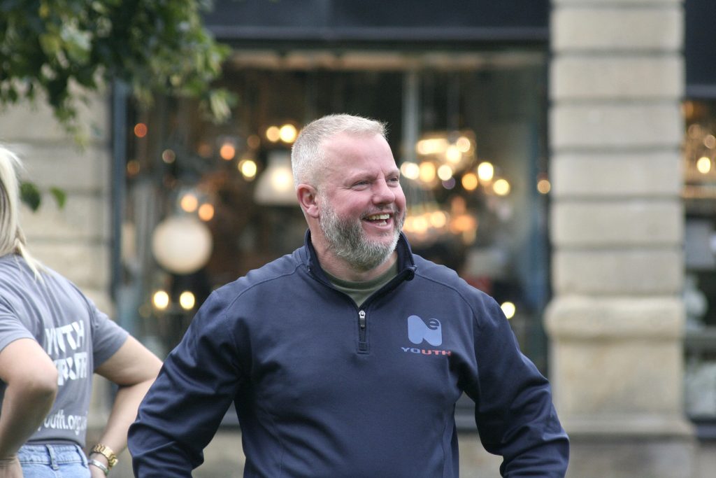 A white male, wearing a blue top smiles at something off camera. He is white, with grey hair and a grey beard.