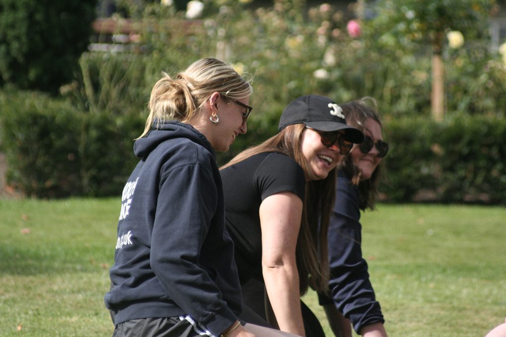 Three white females, sat on grass, laugh together at something unseen.