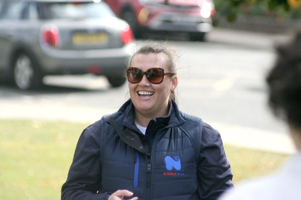 A young white woman with brown hair wearing sunglasses smiles as she looks in the distance.