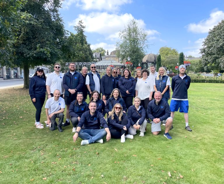 A group of colleagues dressed in company clothing, smile at a camera as their photo is taken. They are on grass with a green backdrop of trees, plants and ornate buildings.